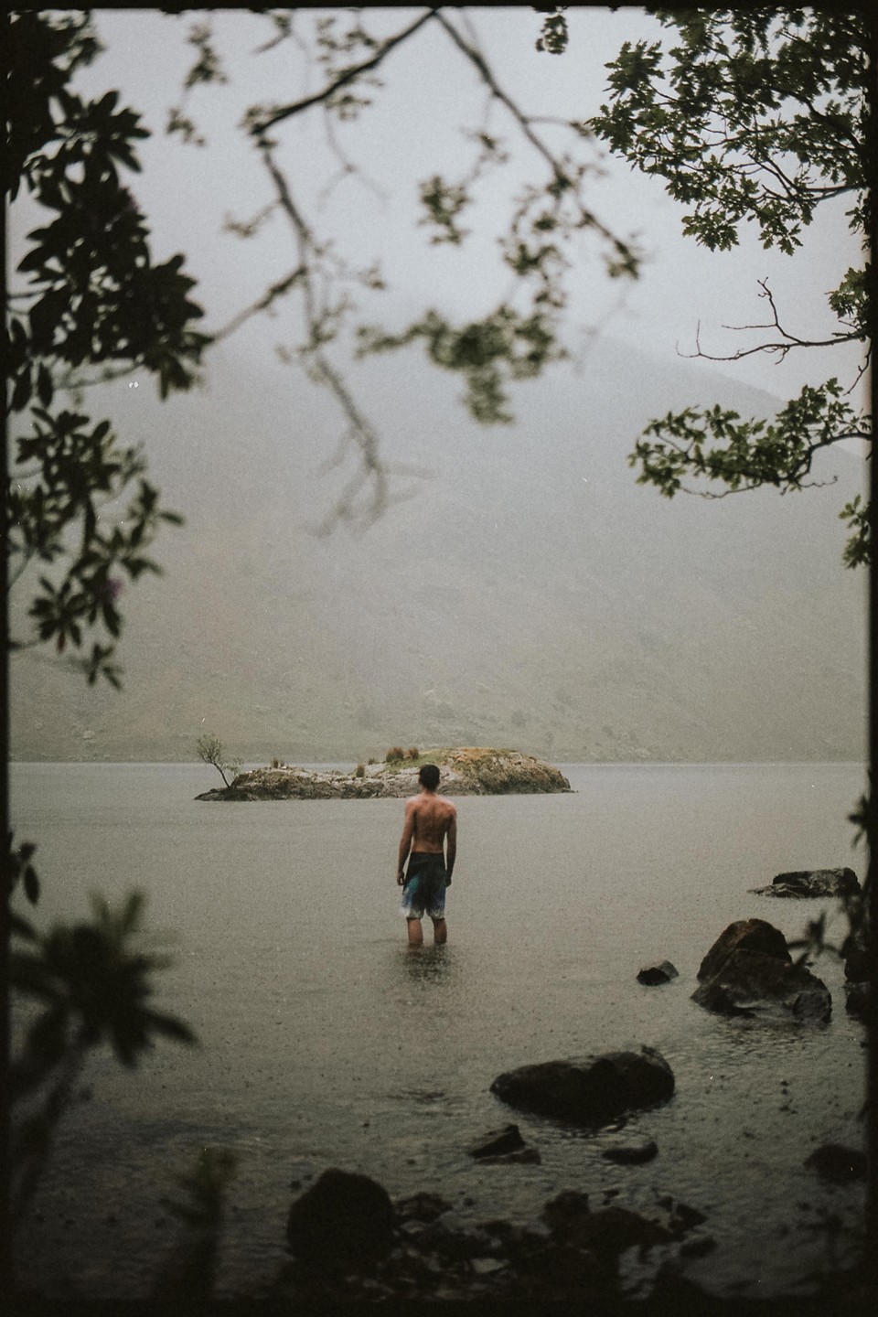 A man stands in a lake with an island in the background, framed by tree branches.