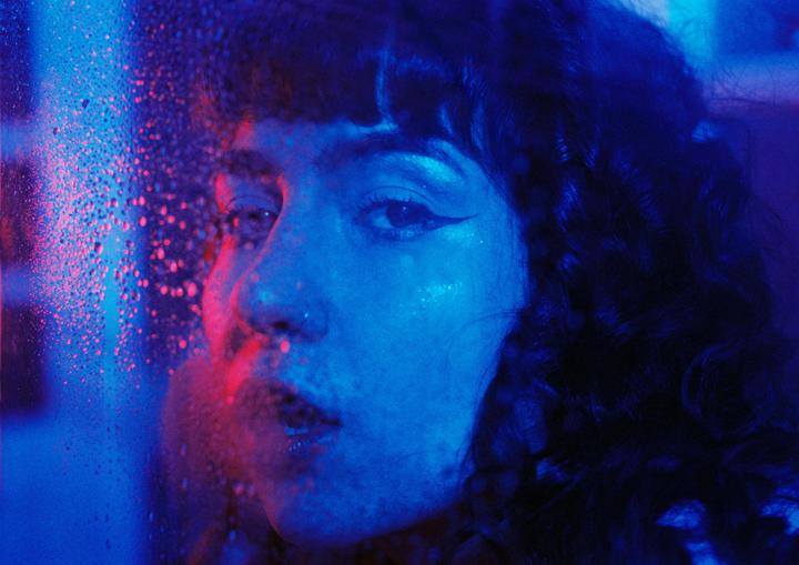 A woman with dark curly hair looks through a wet window, illuminated by blue and red neon light.