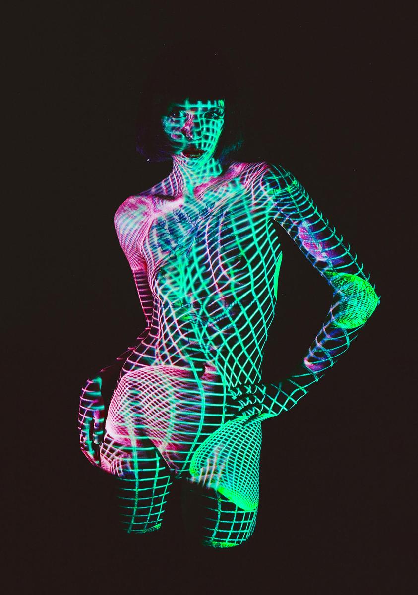 A woman is covered in projected neon green and pink grid patterns against a dark background.
