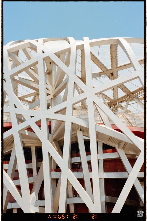 A close-up view of the white, crisscrossing beams of the Beijing National Stadium is shown against a clear blue sky.