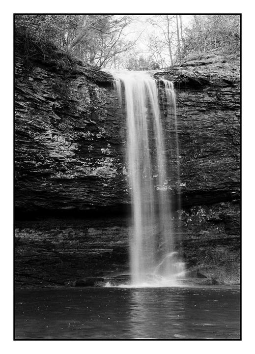 A tall waterfall cascades into a pool of water surrounded by rocky cliffs and trees.