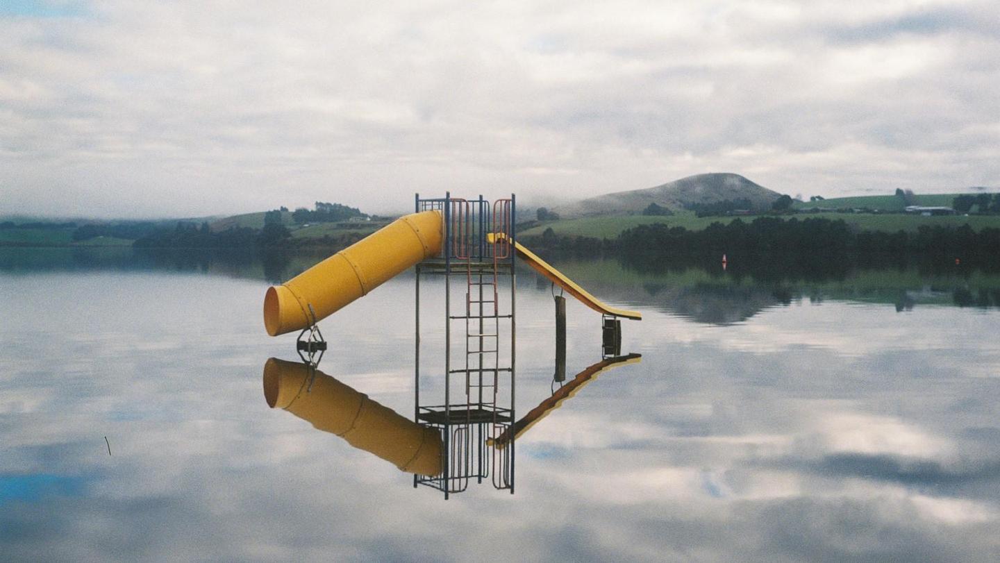 A yellow playground slide structure is partially submerged in calm water, reflecting in the surface, with green hills and a cloudy sky in the background.