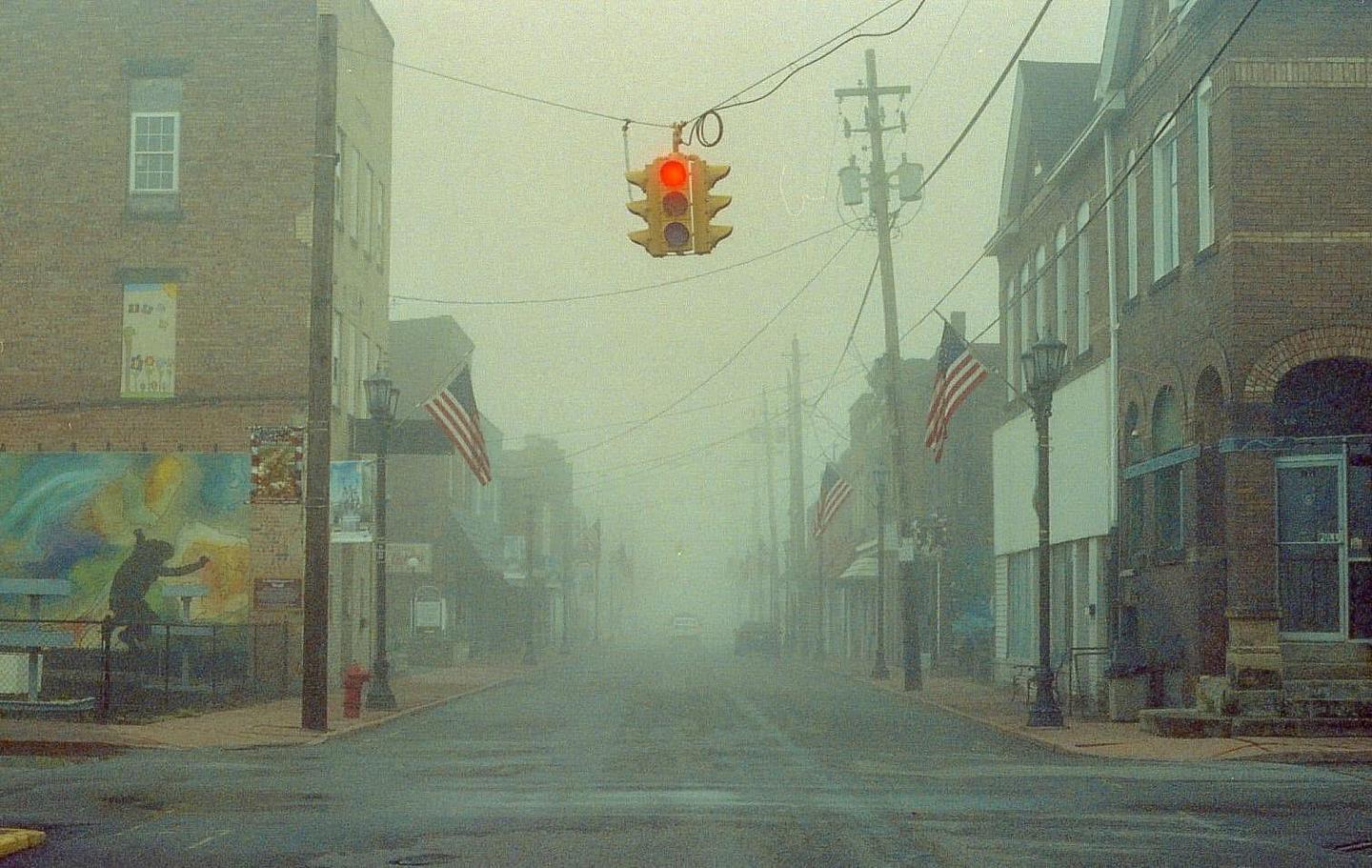 A street with buildings and American flags is shrouded in thick fog, with a red traffic light hanging overhead.
