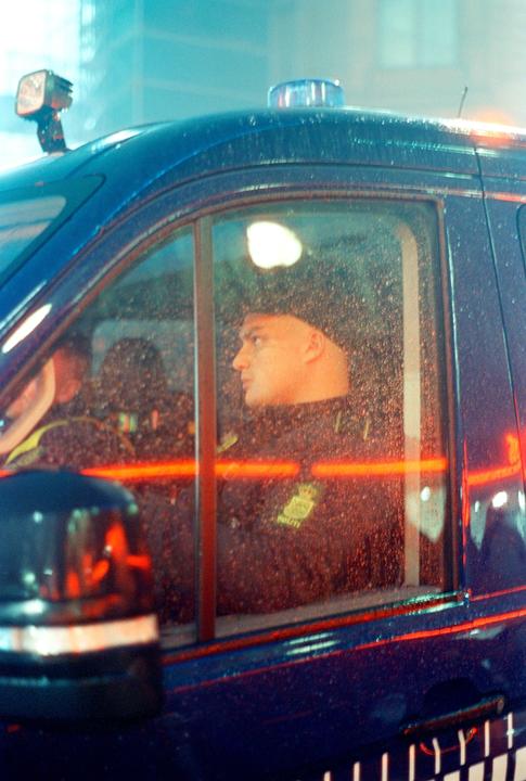 A man in uniform looks out the window of a police vehicle at night.