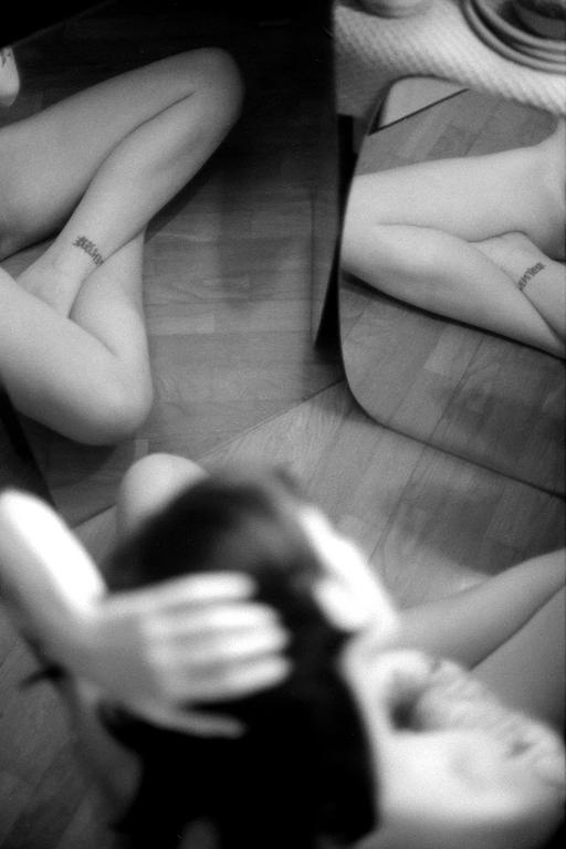 A woman's legs with a tattoo on her ankle are reflected in a mirror, with her head and arms out of focus in the foreground.