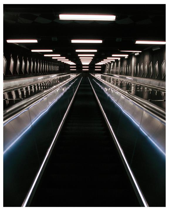 A long, dark escalator tunnel is illuminated by rows of fluorescent lights on the ceiling and glowing handrails.