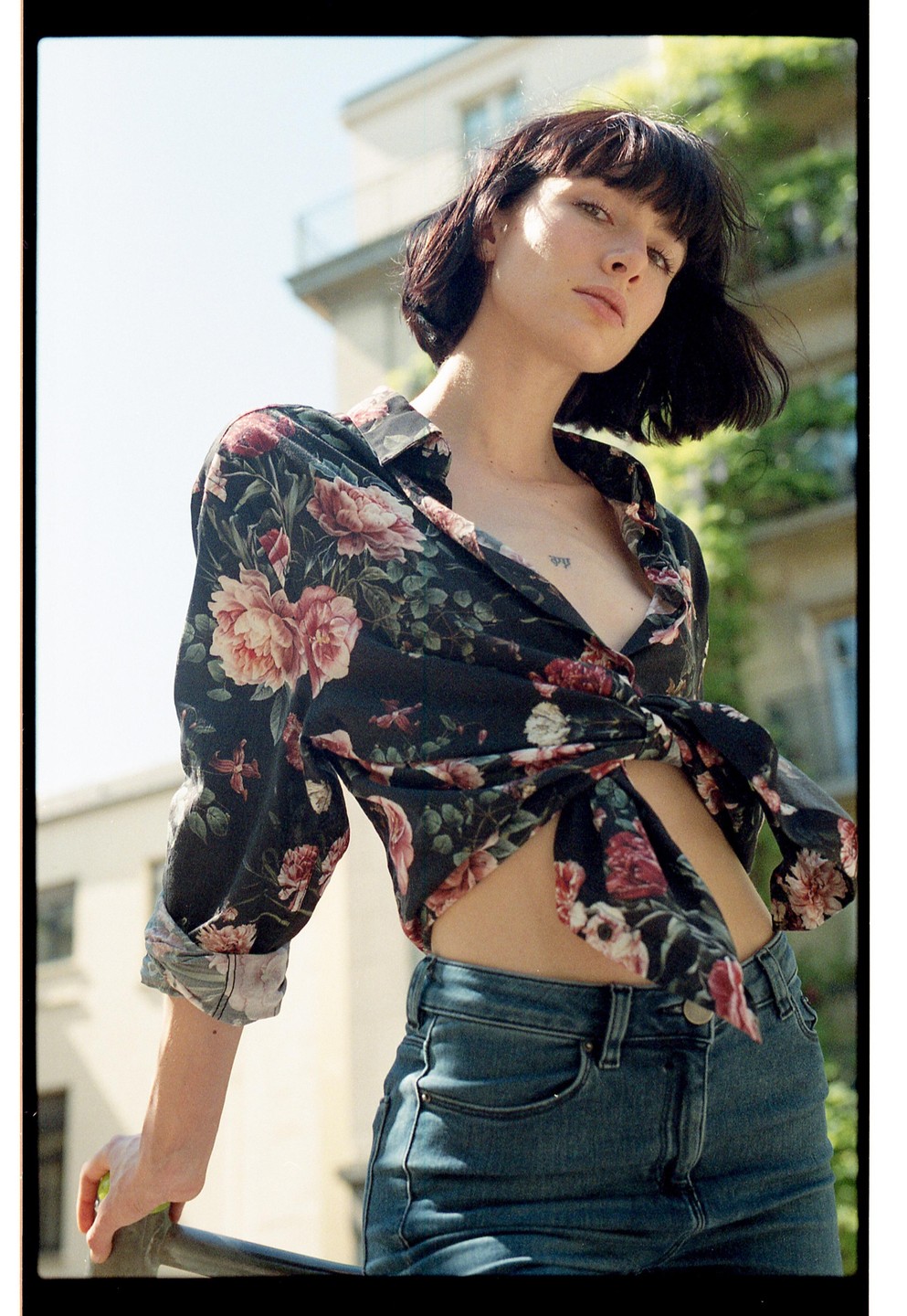 A woman with short dark hair and a floral shirt tied at the waist poses outdoors.