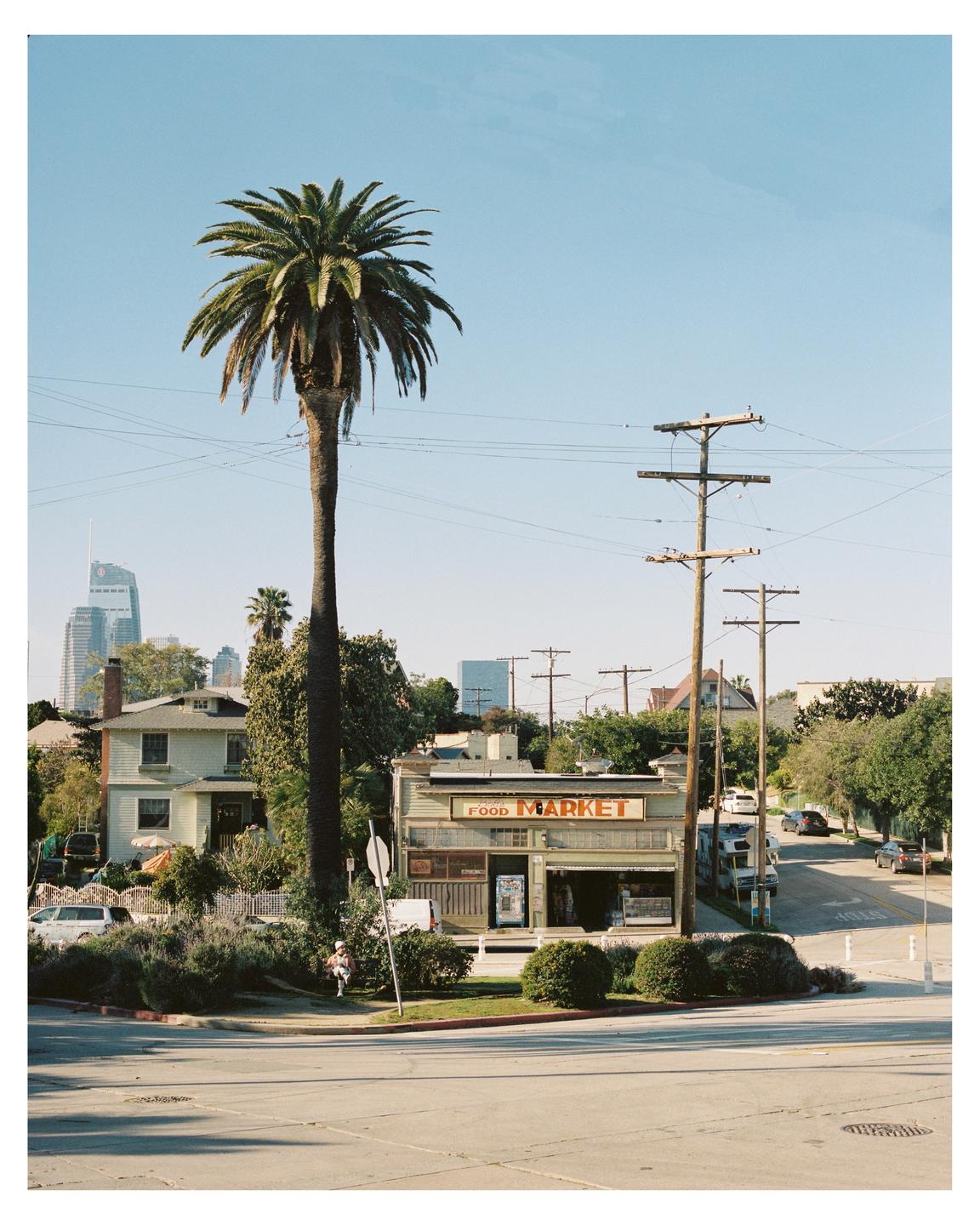 A tall palm tree stands next to a food market on a sunny street corner with buildings and power lines in the background.