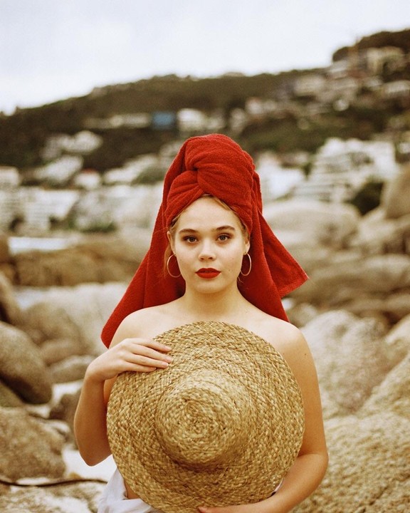 A woman with a red towel wrapped around her head holds a straw hat in front of a rocky, mountainous background.