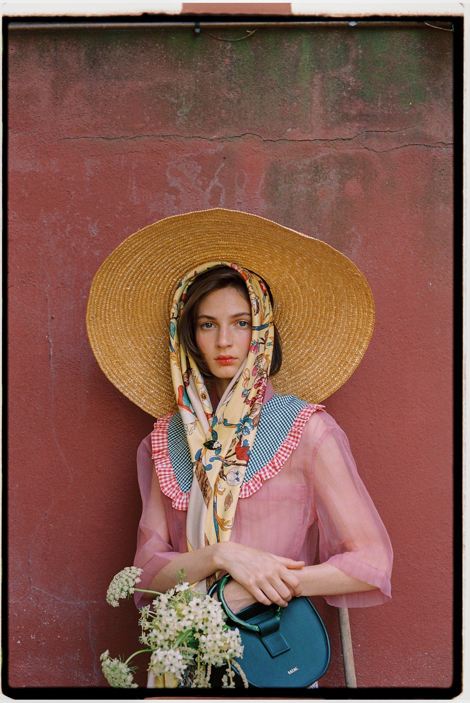 A woman wearing a large straw hat and a patterned scarf stands against a red wall, holding a small handbag and a bouquet of white flowers.
