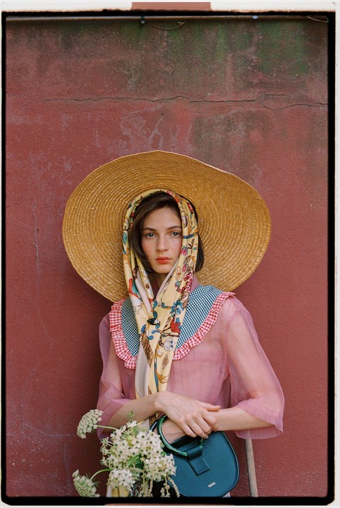 A woman wearing a large straw hat and a patterned scarf stands against a red wall, holding a small handbag and a bouquet of white flowers.