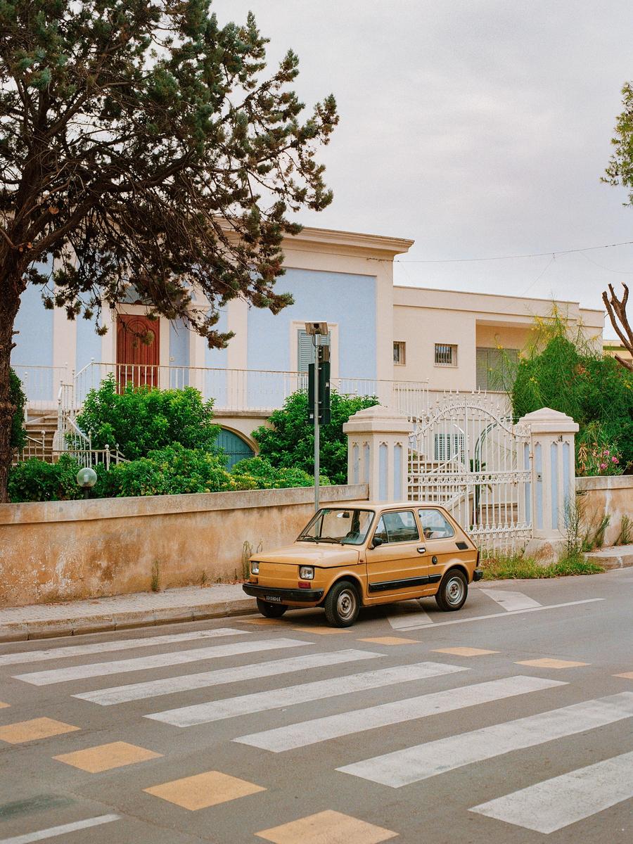 A small yellow car is parked on the side of a street with a crosswalk in the foreground and a building with a blue facade in the background.