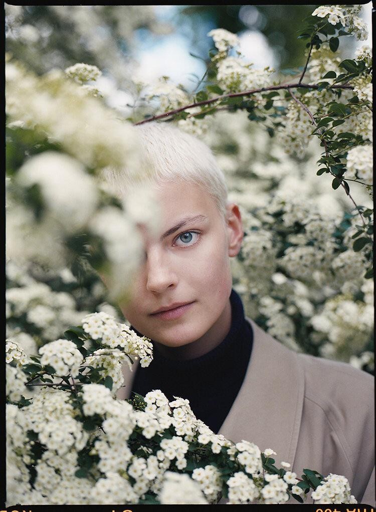 A woman with short blonde hair and blue eyes looks at the viewer through a bush of white flowers.