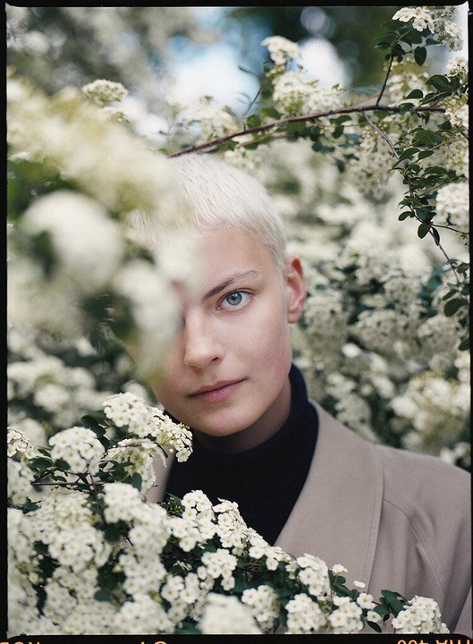 A woman with short blonde hair and blue eyes looks at the viewer through a bush of white flowers.