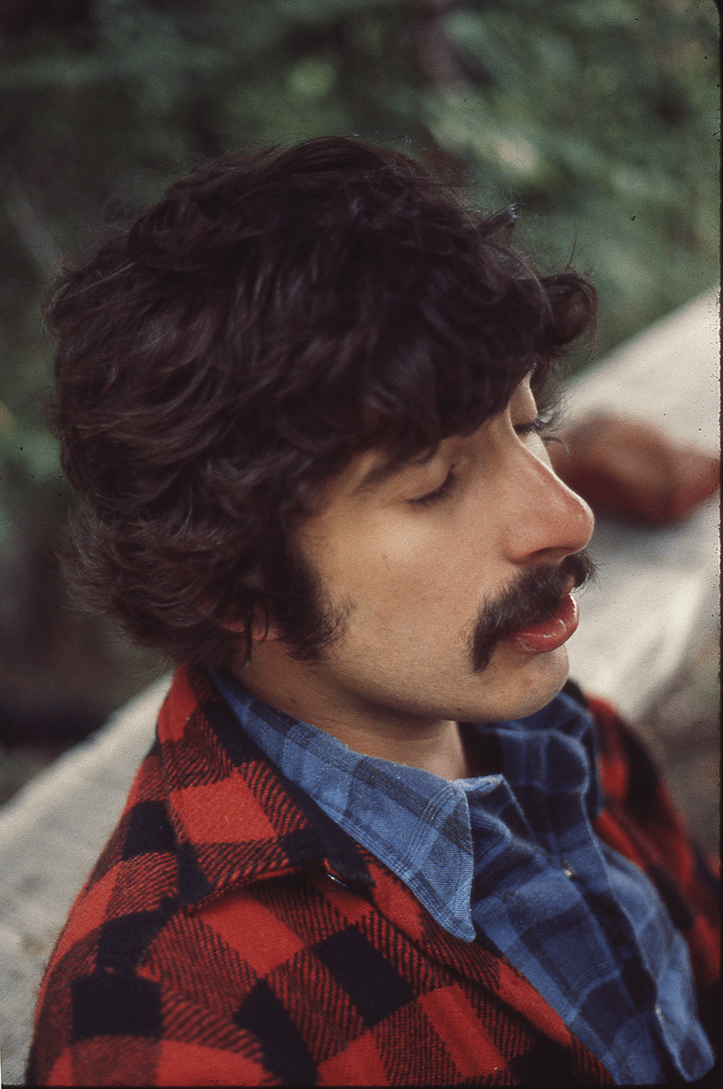 A man with a mustache and curly hair wears a red and black plaid jacket over a blue plaid shirt.