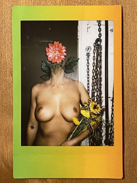 A person with a flower for a head holds a bouquet of sunflowers while standing in front of chains.