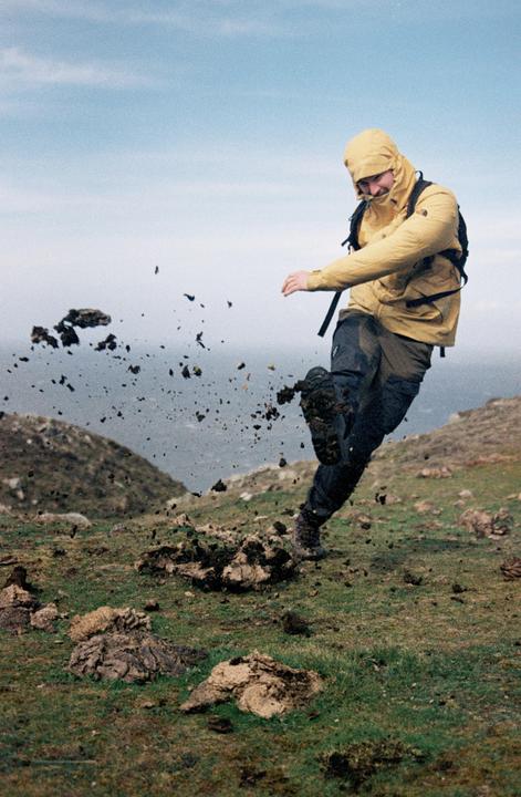 A man in a yellow jacket kicks up dirt and mud on a grassy hillside.