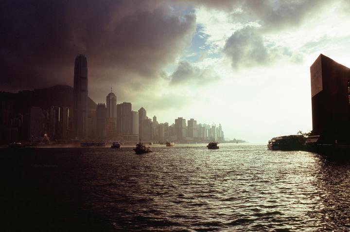 A cityscape with tall buildings is visible across a body of water with several boats, under a dramatic cloudy sky.
