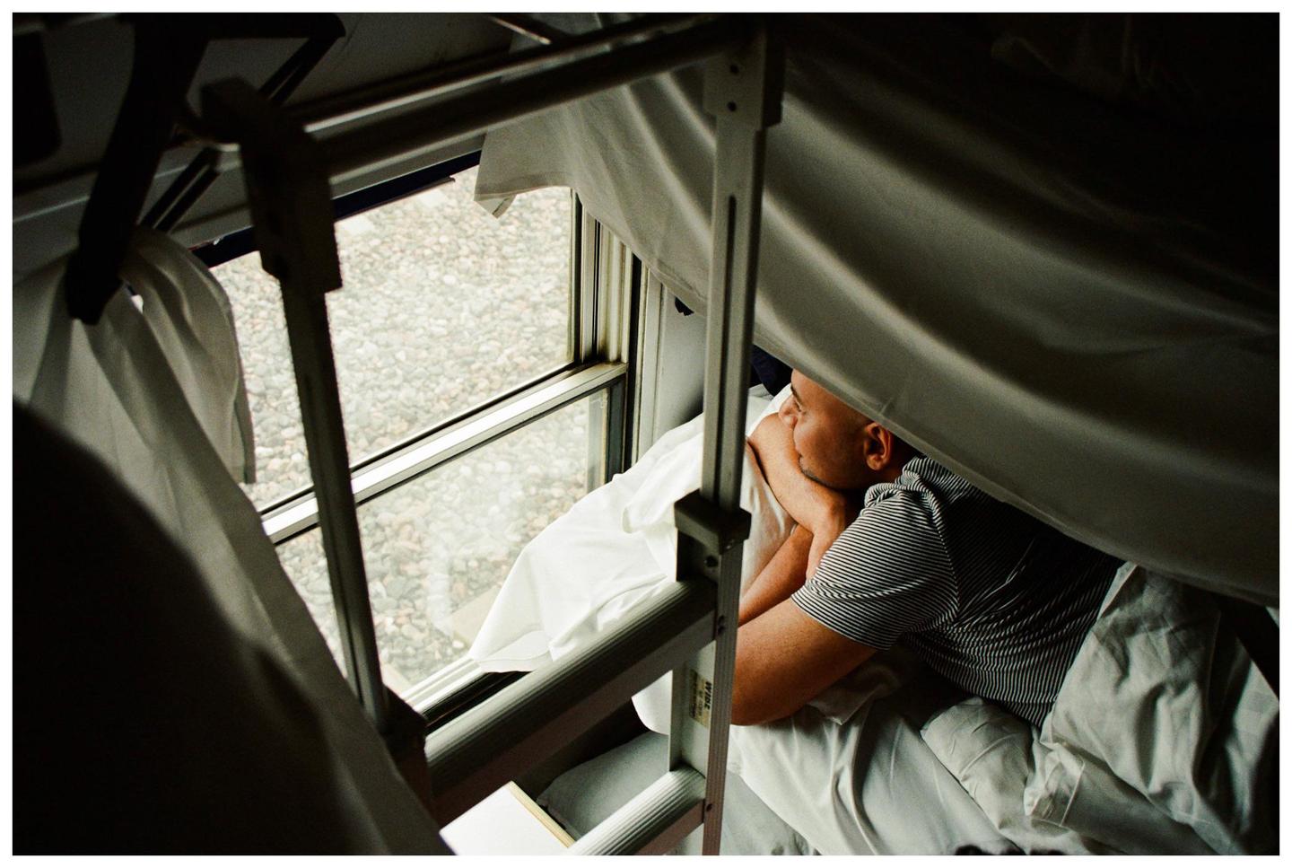 A man lies in a bunk bed on a train, looking out the window.