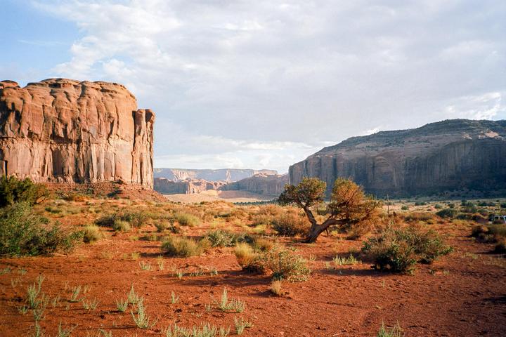 A desert landscape features red earth, green shrubs, and large rock formations under a cloudy sky.