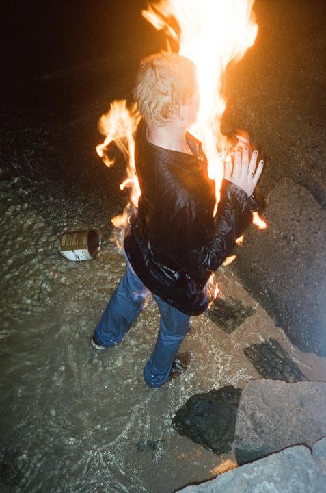 A person stands in shallow water with flames rising around their head and shoulders.