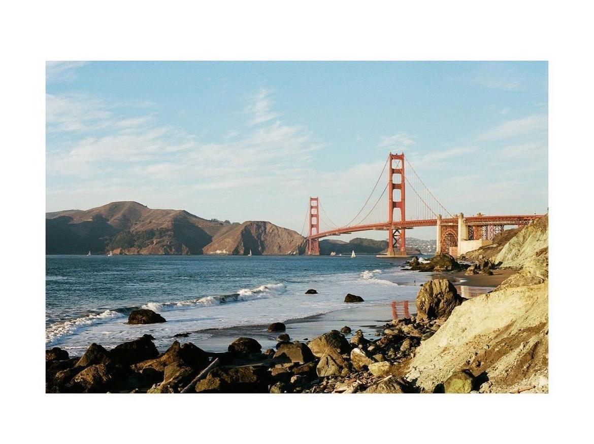 The Golden Gate Bridge spans across a body of water with waves crashing on a rocky beach in the foreground and hills in the background.