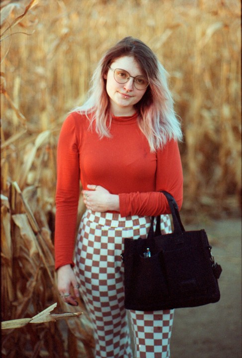 A woman with glasses and a nose ring stands in a field of dry corn stalks, holding a black bag.