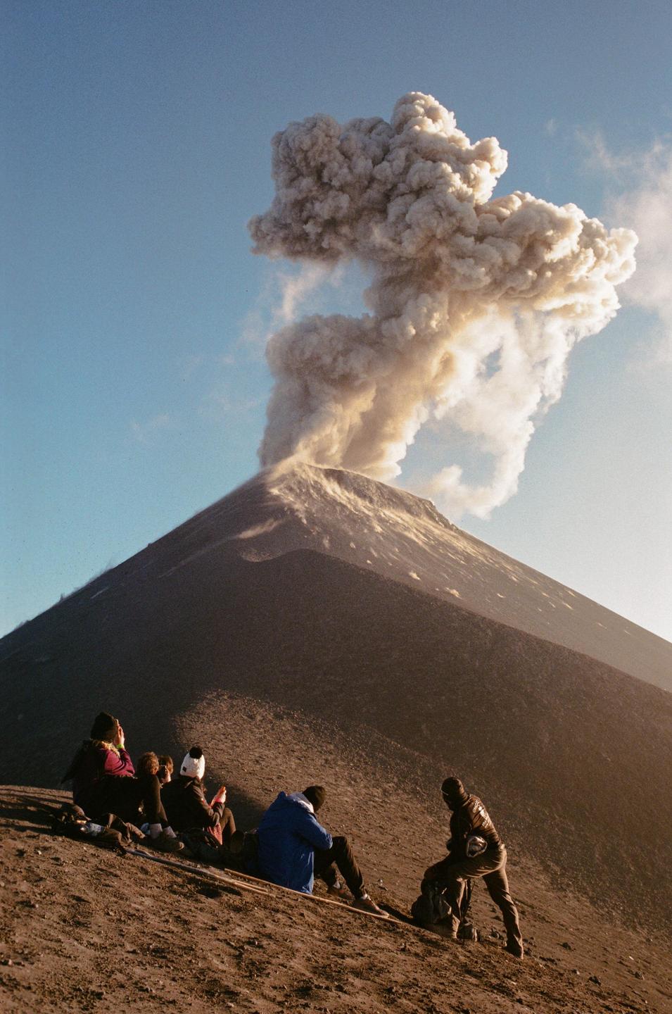 A group of people sit on a volcanic slope watching a volcano erupt.