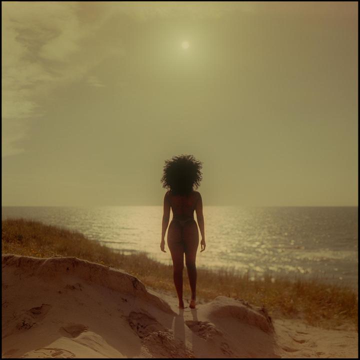 A woman with curly hair stands on a sandy dune looking out at the ocean under a hazy sun.