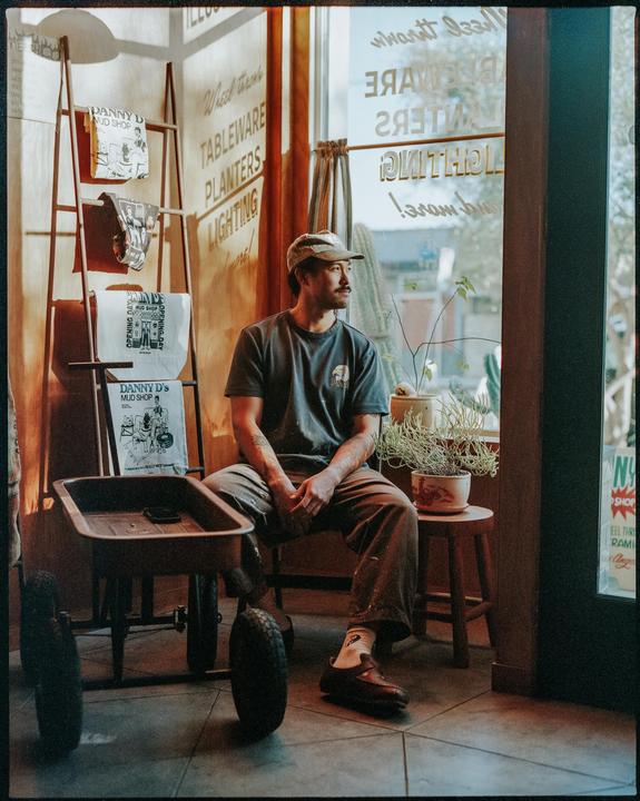 A man with a mustache and cap sits in a workshop next to a red wagon and potted plants, looking out a window.