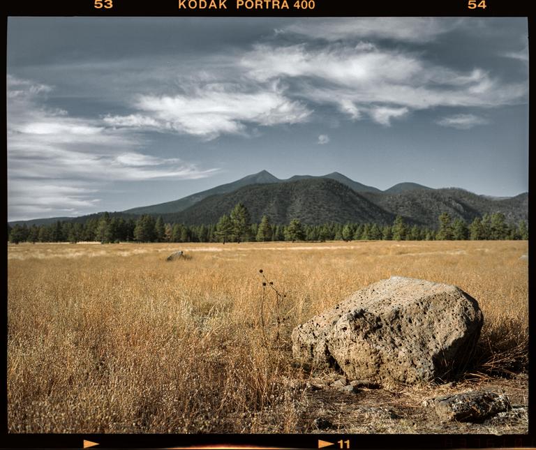 A large rock sits in a field of dry grass with mountains and a forest in the background under a cloudy sky.