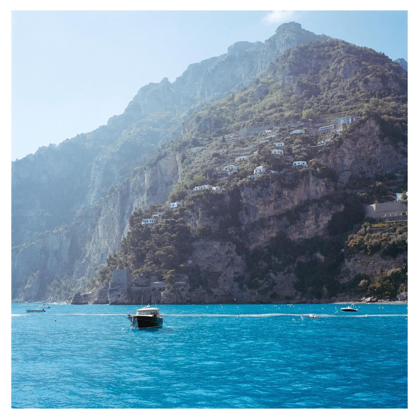A boat floats on bright blue water in front of a steep, rocky coastline dotted with white buildings.