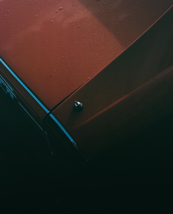 Raindrops cover the orange hood of a car, with a blue stripe visible on the side.