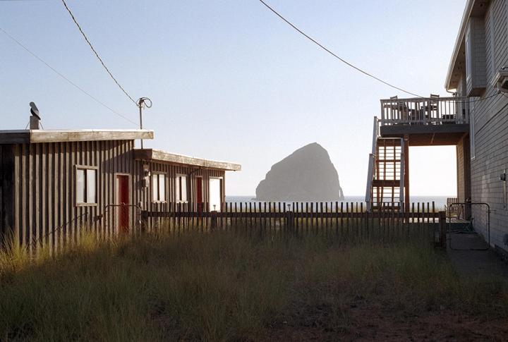 A large rock formation rises from the ocean behind a wooden fence and two buildings.