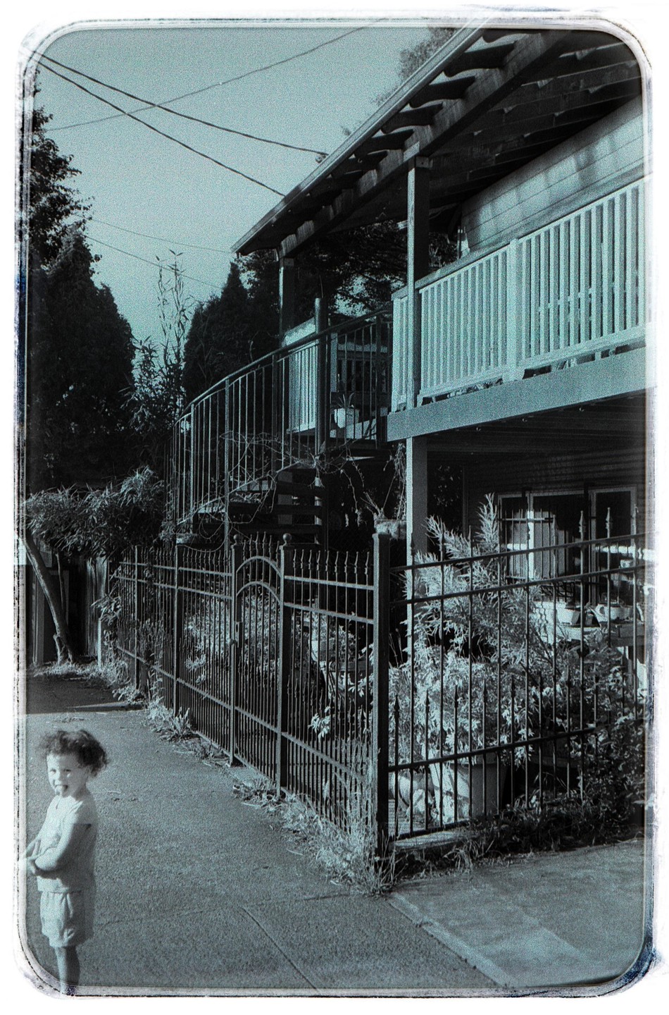 A child stands on a sidewalk next to a metal fence and a house with a balcony.