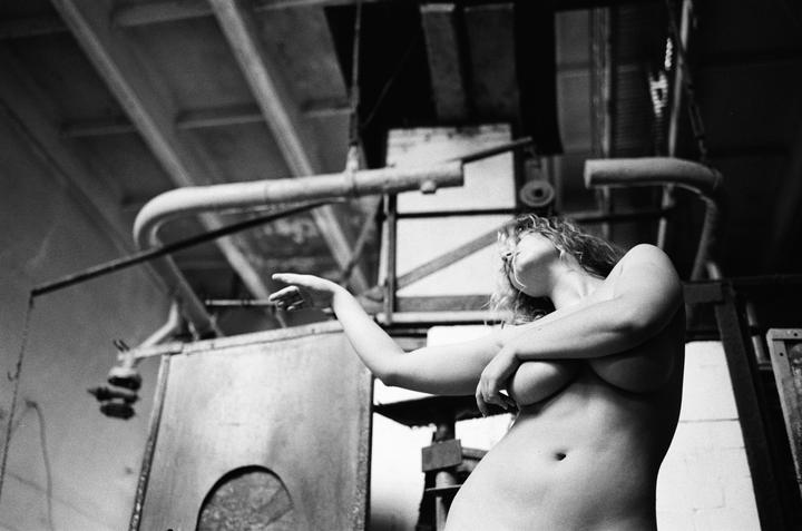 A topless woman with curly hair stands in an industrial setting, looking up and pointing to the left.