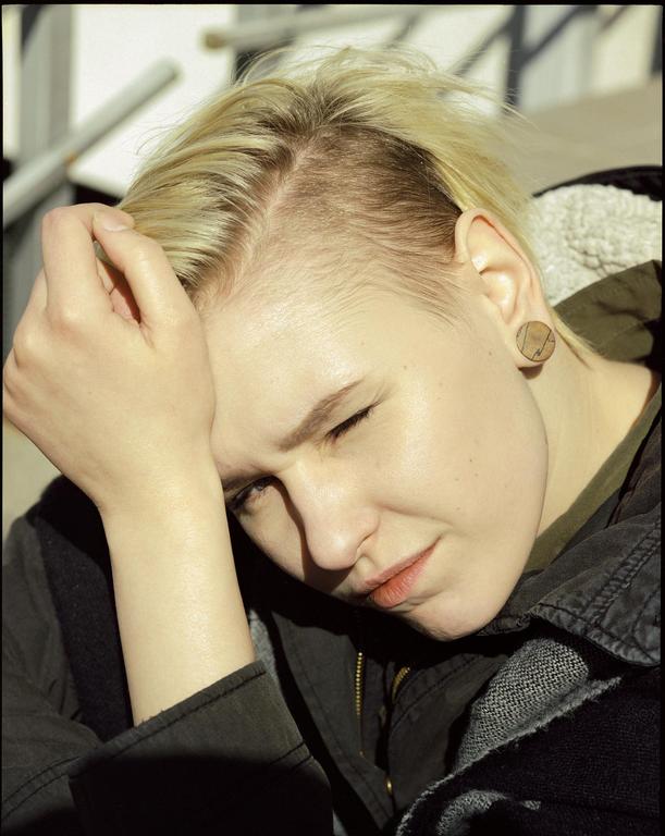 A woman with short blonde hair and a dark jacket rests her head on her arm, with her eyes closed and face partially in shadow.