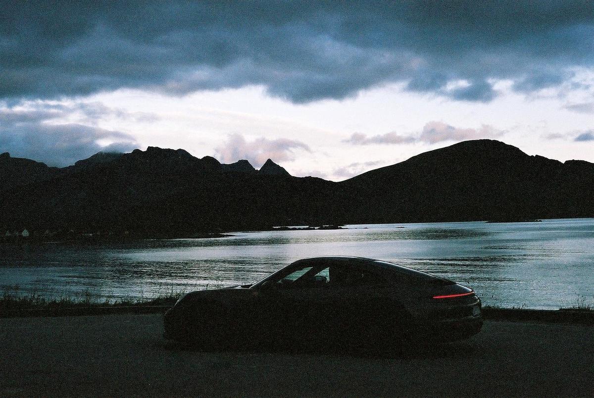 A dark car is parked on a road overlooking a body of water with mountains in the background under a cloudy sky.