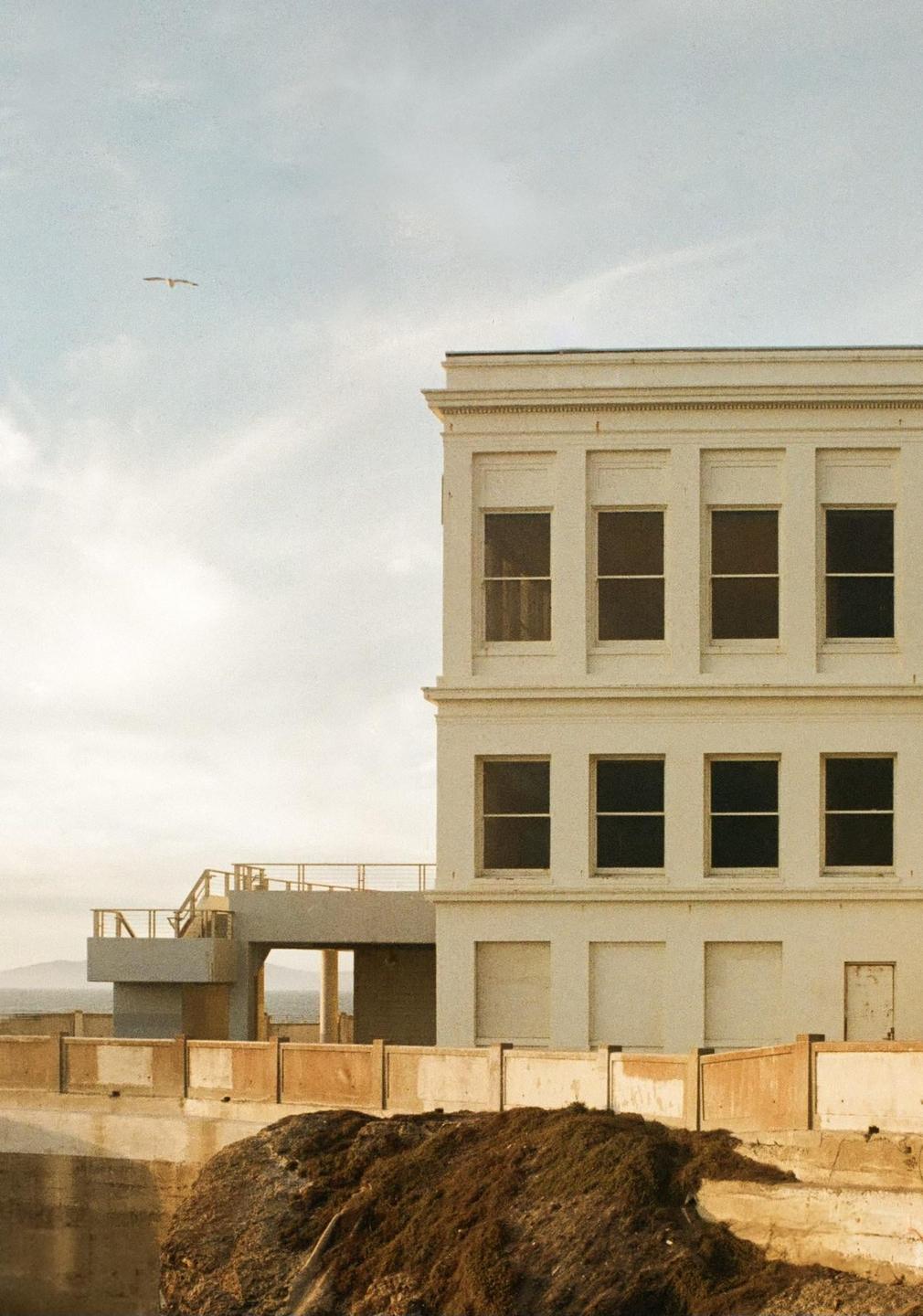 A white building with multiple windows stands on a cliff overlooking the ocean, with a bird flying in the sky.