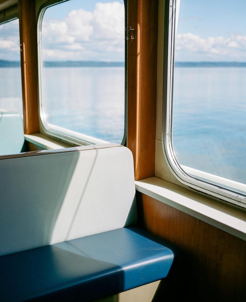A blue and white seat is visible inside a ferry, with a window looking out onto a body of water and a cloudy sky.