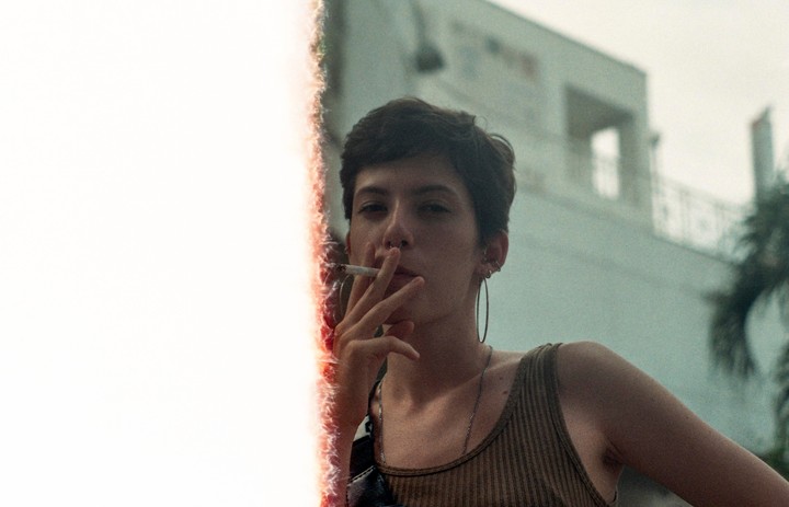 A woman with short hair and a nose ring holds a lit cigarette to her mouth, with a bright light leak on the left side of the frame.