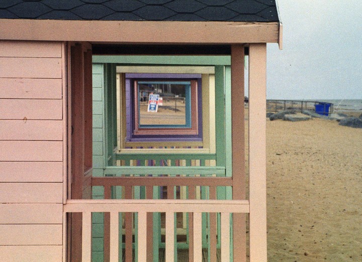 A series of pastel-colored beach hut frames create a tunnel effect looking out onto a sandy beach.