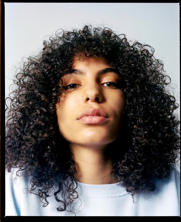 A woman with curly hair looks directly at the viewer.