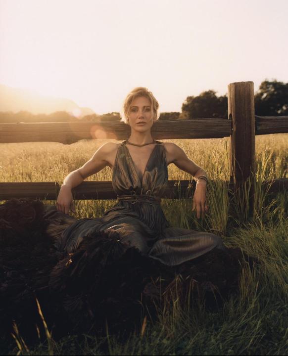 A woman in a dress sits on a dark blanket in a field with a wooden fence behind her at sunset.