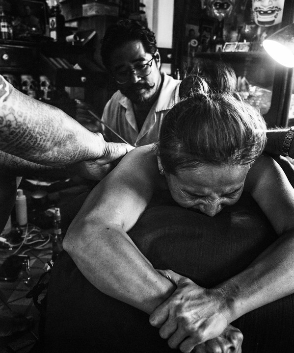 A woman grimaces in pain while receiving a tattoo from a man with a mustache and glasses.