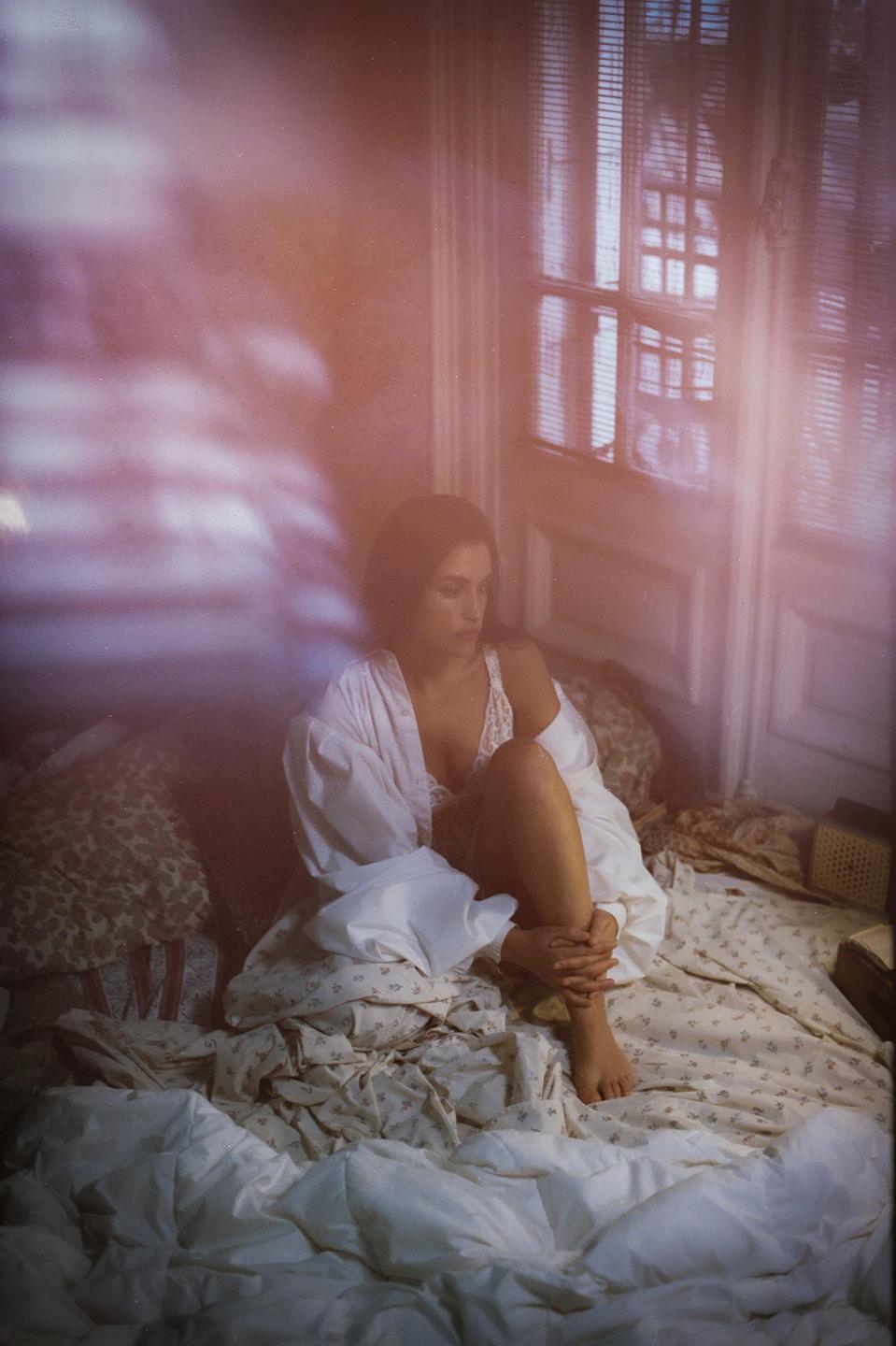 A woman sits on a bed in a sunlit room, looking down at her hands.