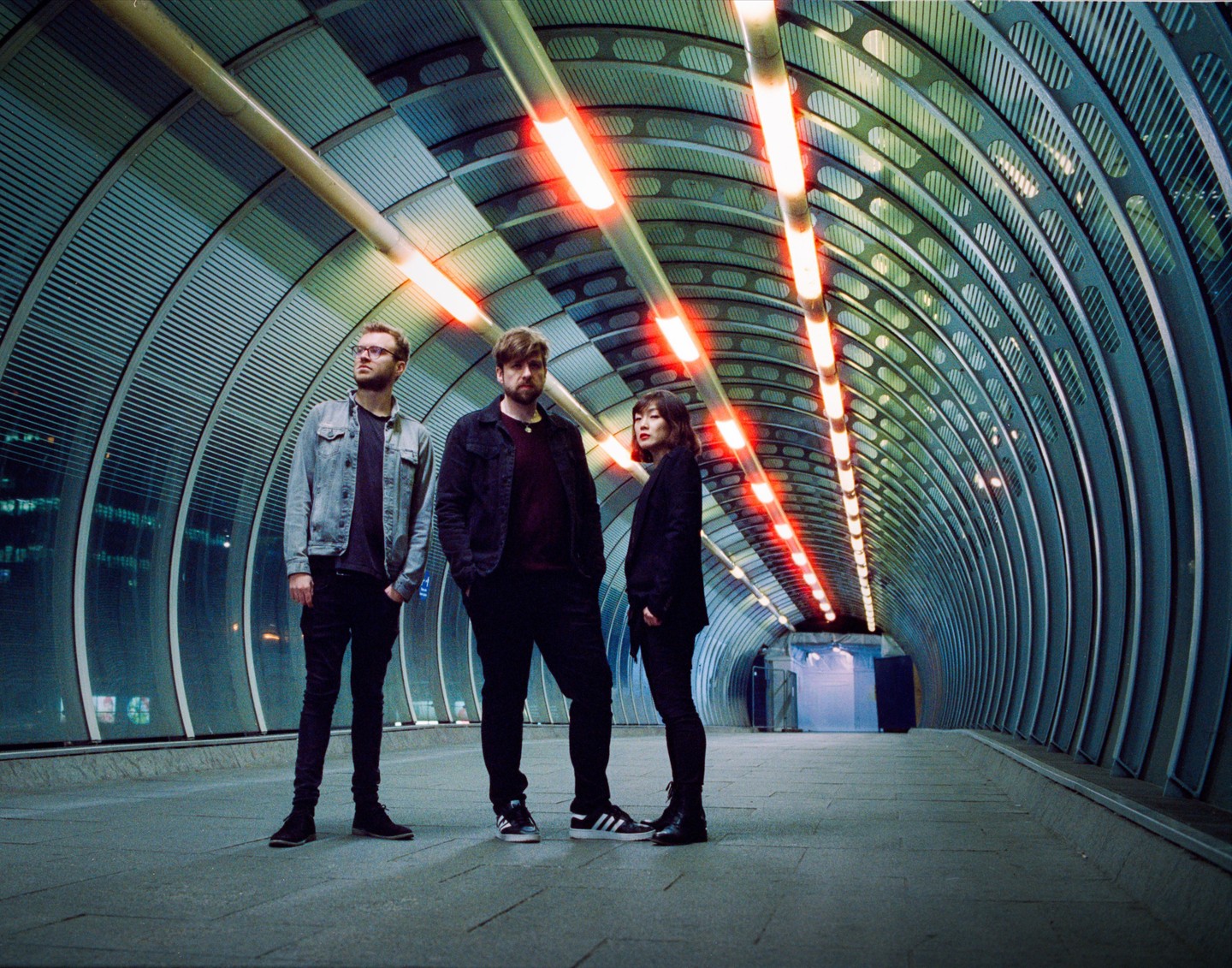 Three people stand in a futuristic tunnel with red lights on the ceiling.