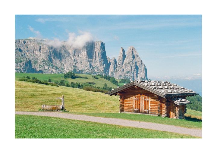 A wooden cabin with a stone roof sits on a grassy hill with a mountain range in the background.