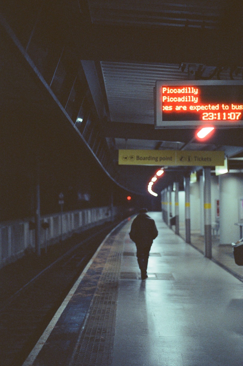 A person walks away from the camera on a subway platform at night, with a red digital sign displaying "Piccadilly" and the time "23:11:07" in the background.