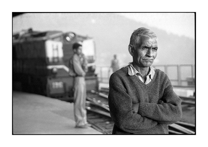 A man with a mustache and folded arms stands on a train platform with a train and another man in the background.